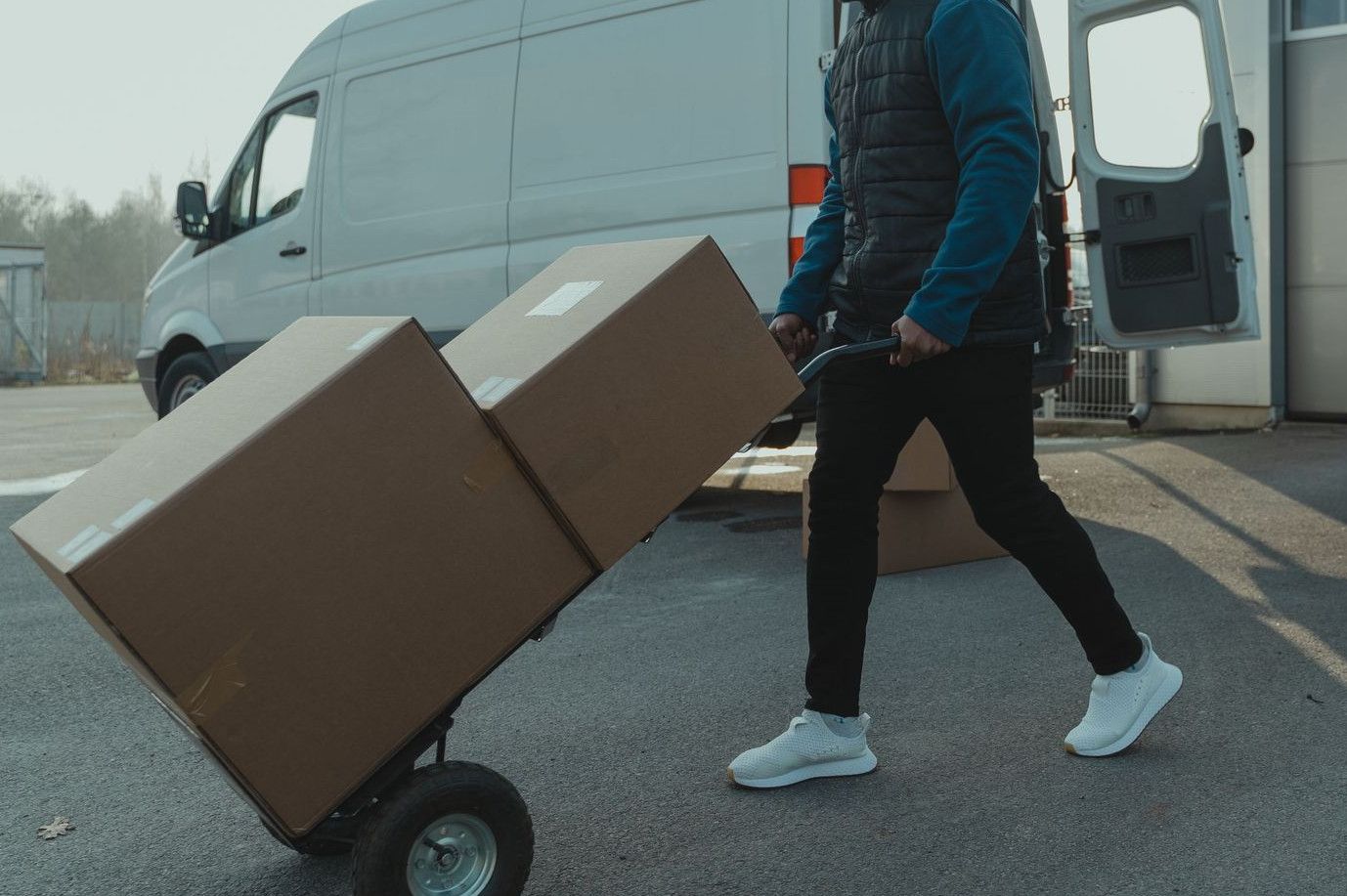 Courier pushing a trolley with boxes near a van at a warehouse, showcasing delivery services.